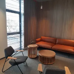 Nowoczesne wnętrze biurowe z pomarańczową skórzaną sofą, stolikami kawowymi z drewnianymi lamelami i szarymi fotelami na metalowych nogach, oświetlone światłem dziennym wpadającym przez żaluzje.