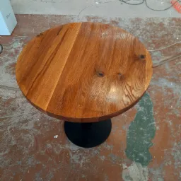 Okrągły, drewniany st&oacute;ł z wyraźnym usłojeniem, polakierowany na wysoki połysk, osadzony na czarnej, okrągłej podstawie. Widoczny na tle betonowej podłogi z resztkami farby i śladami po pracy.
