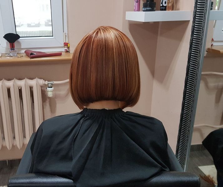 Krótkie, proste bob w odcieniach miedzi z blond refleksami, widok z tyłu, klientka w czarnej pelerynie fryzjerskiej.