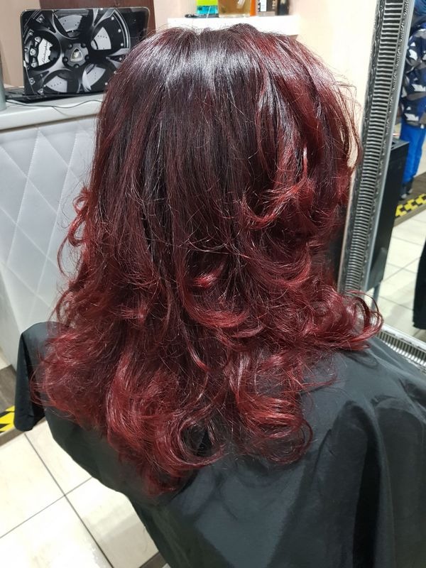 Tył głowy kobiety z długimi, pofalowanymi włosami w odcieniach ciemnej wiśni i mahoniu, świeżo po koloryzacji i stylizacji w salonie, widoczny czarny fartuch ochronny.