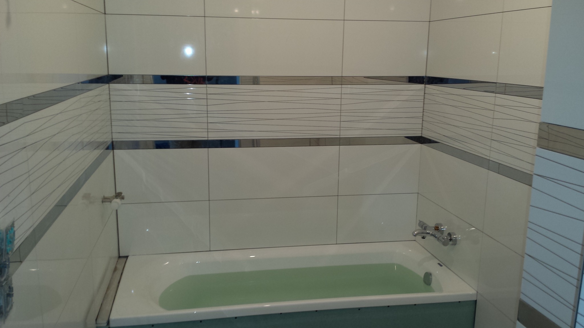 Wykończona łazienka z białą wanną wypełnioną wodą, ściany wyłożone białymi płytkami z dekoracyjnym pasem z cienkimi, falistymi liniami i metalowymi akcentami.