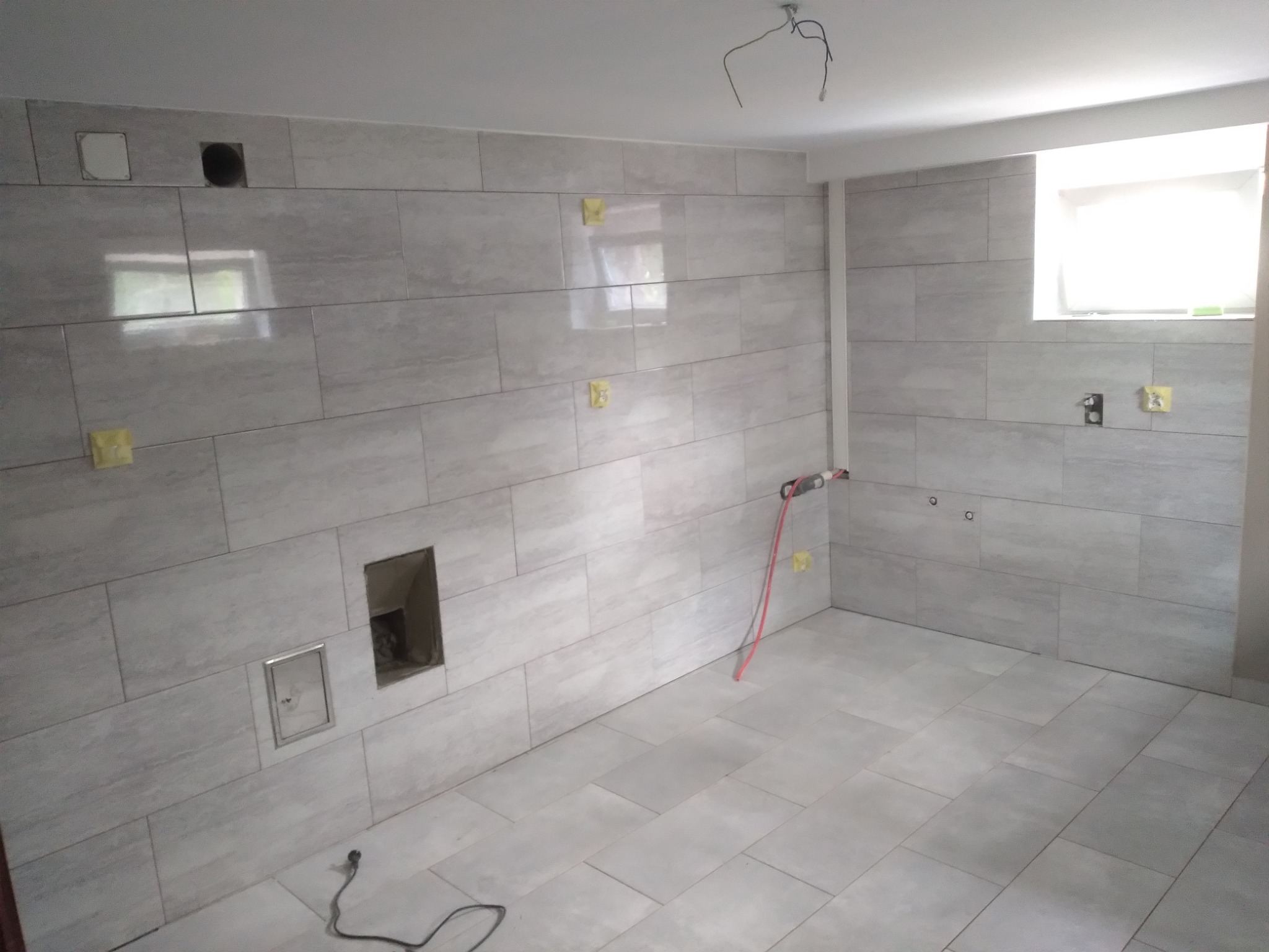 Wnętrze w trakcie prac wykończeniowych, ściany i podłoga pokryte szarymi, prostokątnymi płytkami, widoczne otwory w ścianie, wystające kable, rurka instalacyjna w kolorze czerwonym, żółte taśmy...