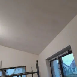 Wnętrze w trakcie prac wykończeniowych: świeżo wygładzony sufit z widocznymi śladami szpachlowania, okno zabezpieczone folią, rusztowanie w rogu pomieszczenia.