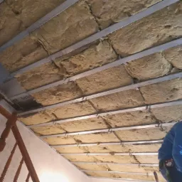 Montaż izolacji z wełny mineralnej na metalowym ruszcie pod sufitem, widoczne elementy drewnianej balustrady schodów w tle.