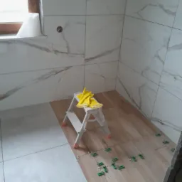 Układanie płytek podłogowych w łazience, widoczne płytki imitujące drewno, krzyżyki dystansowe, drabinka z żółtymi rękawicami roboczymi, ściany wyłożone białymi płytkami z szarym marmurkowym wzorem.