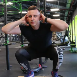 Mężczyzna w czarnym stroju sportowym ćwiczy przysiady na drewnianej skrzyni w siłowni, z rękami splecionymi za głową i wzrokiem skierowanym w dal.