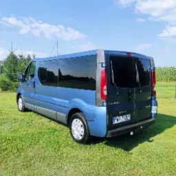 Niebieski bus Renault Trafic z przyciemnianymi szybami, zaparkowany na trawniku w słoneczny dzień, widok z tyłu i boku, z widoczną tablicą rejestracyjną.