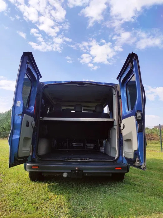 Niebieski van z otwartymi drzwiami bagażnika, widok z tyłu na trawniku w słoneczny dzień.