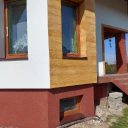 Fragment elewacji domu z oknami, gdzie część ściany wykończona jest imitacją drewna, a cokół budynku ma czerwoną, ziarnistą strukturę.