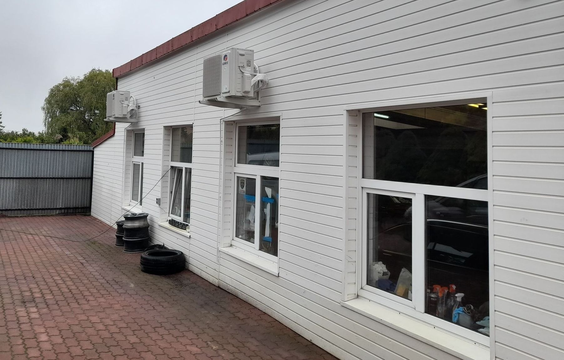 Biały budynek z trzema klimatyzatorami typu split zamontowanymi na elewacji, widoczne opony i felgi samochodowe, kostka brukowa na podwórku.