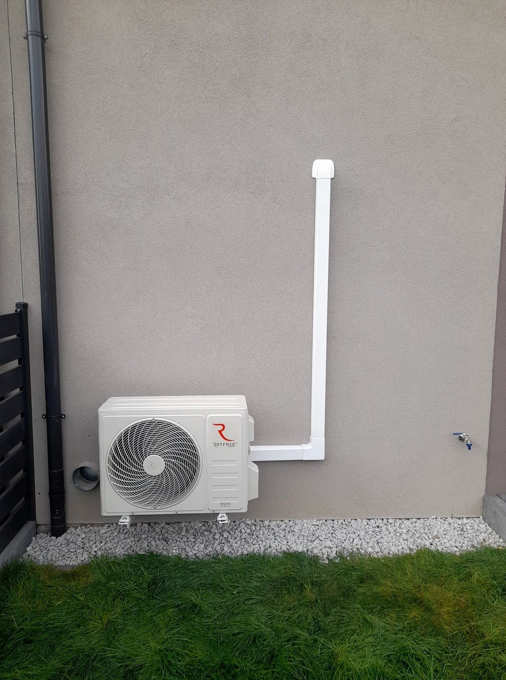 Biała jednostka zewnętrzna klimatyzacji Rotenso zamontowana na szarej ścianie budynku, z widocznym odprowadzeniem skroplin wykonanym z białych rur PCV, podsypana białym gresem, z fragmentem...