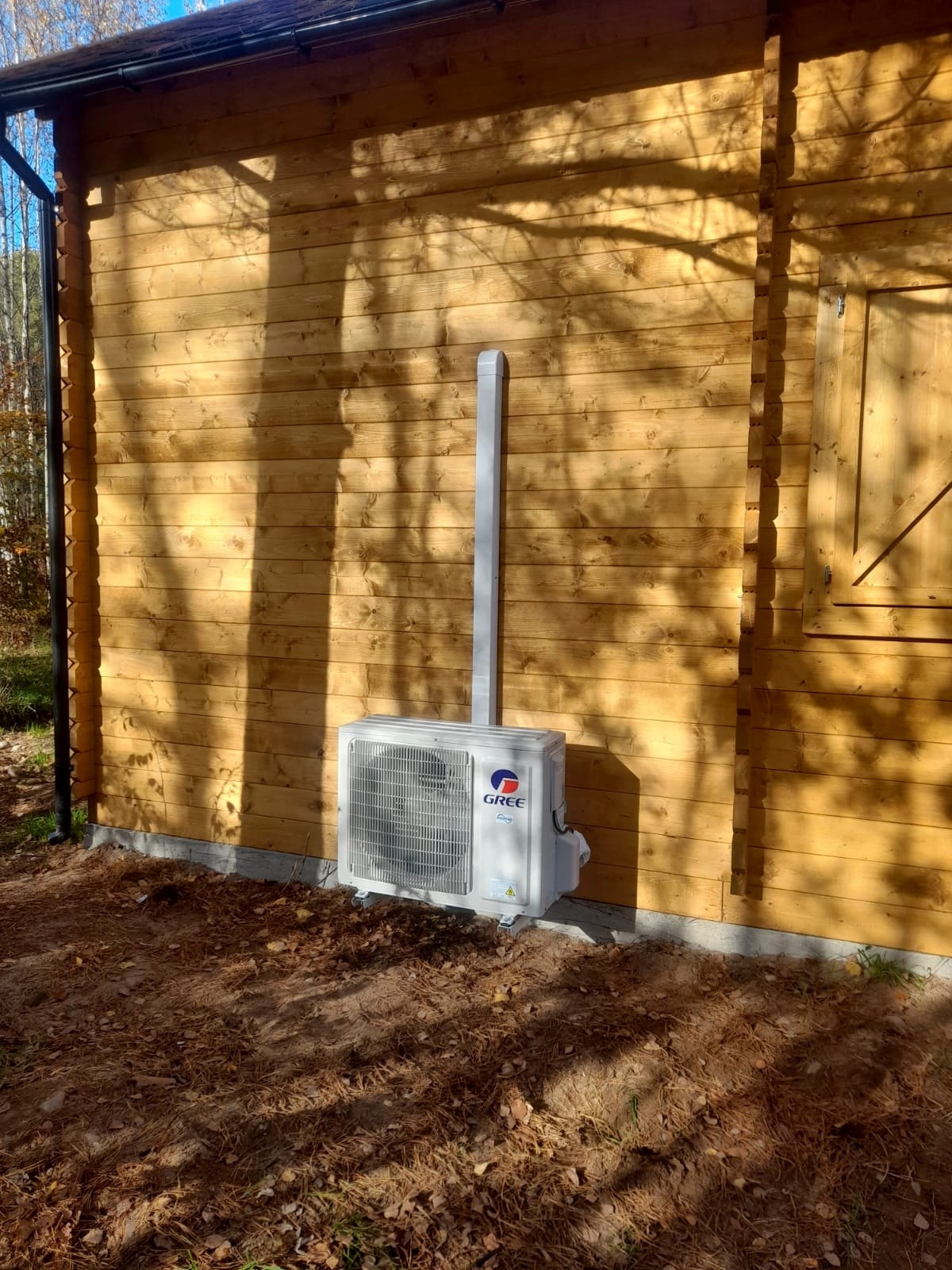 Biała jednostka zewnętrzna klimatyzacji marki Gree zamontowana na drewnianej ścianie domku letniskowego, z widoczną rurą odprowadzającą skropliny, oświetlona popołudniowym słońcem rzucającym cienie...