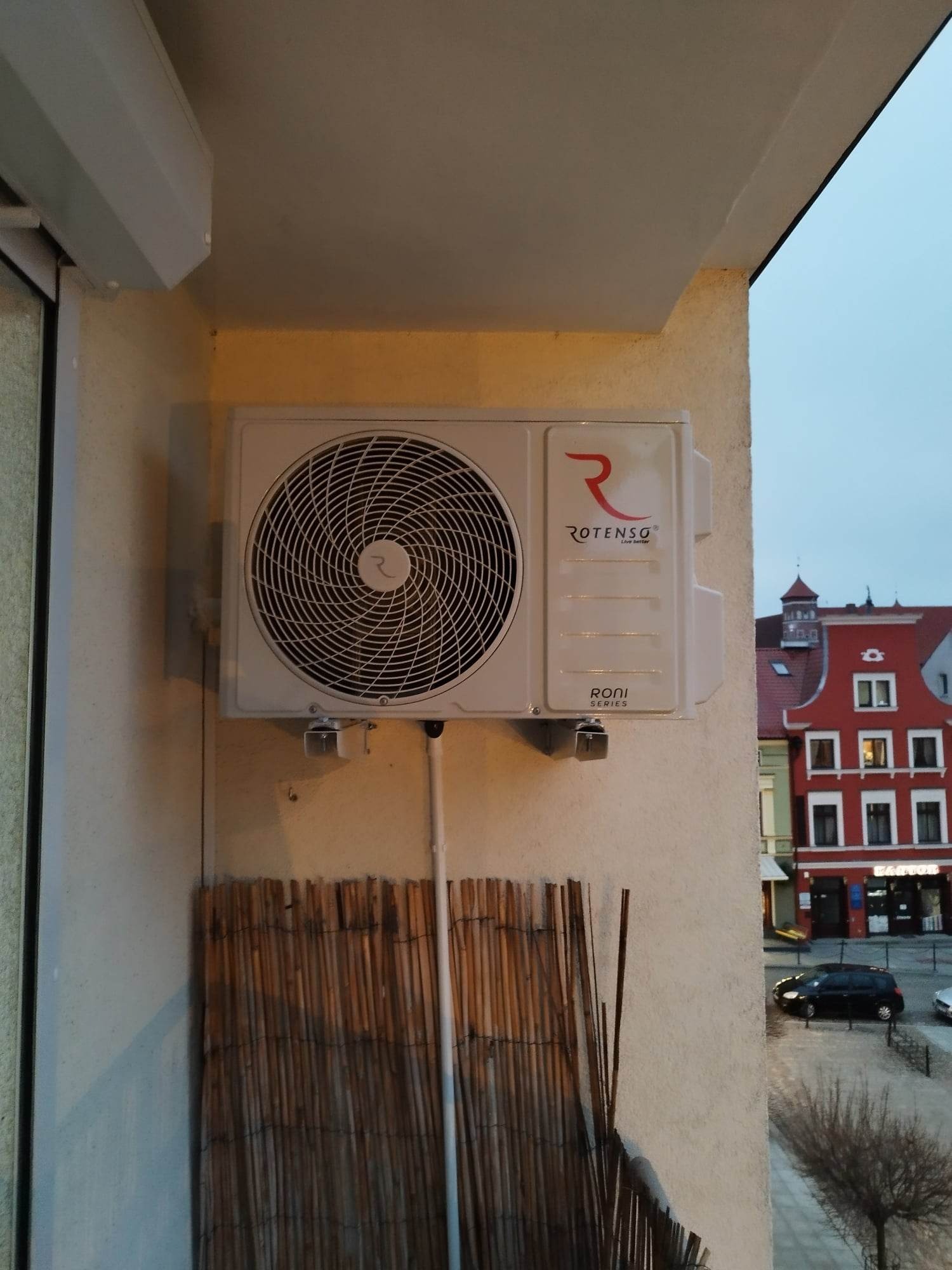 Zewnętrzna jednostka klimatyzacji Rotenso zamontowana na balkonie z widokiem na budynek w tle.