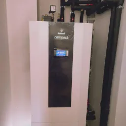 Biała jednostka Galmet Compact z wyświetlaczem, podłączona do czarnych rur instalacji grzewczej, zamontowana przy ścianie.