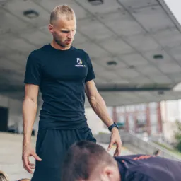 Młody mężczyzna w czarnym sportowym stroju z logo firmy na koszulce obserwuje ćwiczących ludzi na schodach miejskich.