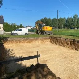 Wykop pod fundamenty domu jednorodzinnego, widoczna żółta ziemia, zabezpieczenie wykopu drewnianą belką, na górze koparka Atlas 1404 i biały samochód dostawczy na tle zieleni i błękitnego nieba.