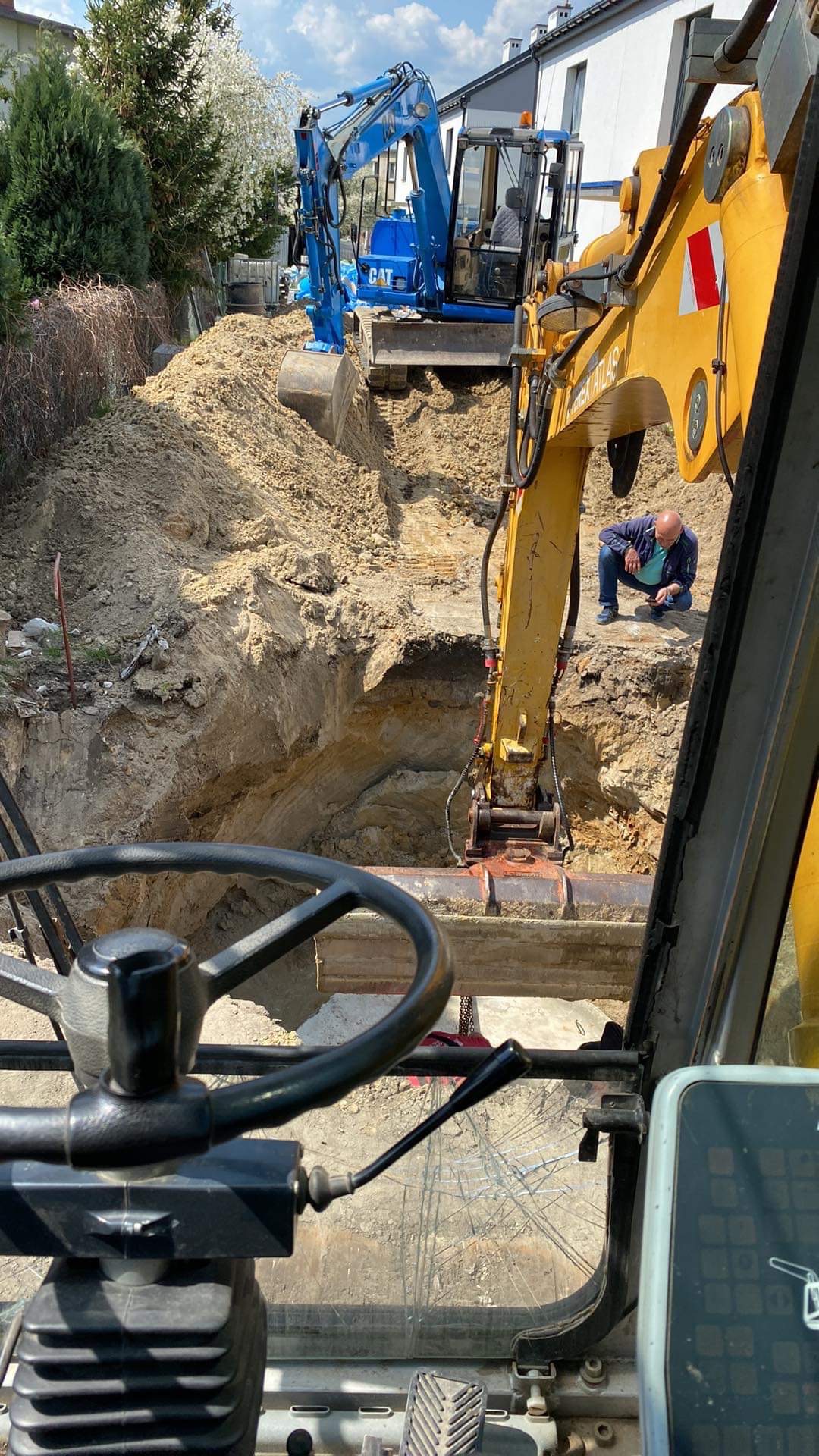 Widok z kabiny koparki na wykop w ziemi, z niebieską koparką CAT w tle i mężczyzną przyglądającym się wykopowi, szyba kabiny z pajęczyną pęknięć.