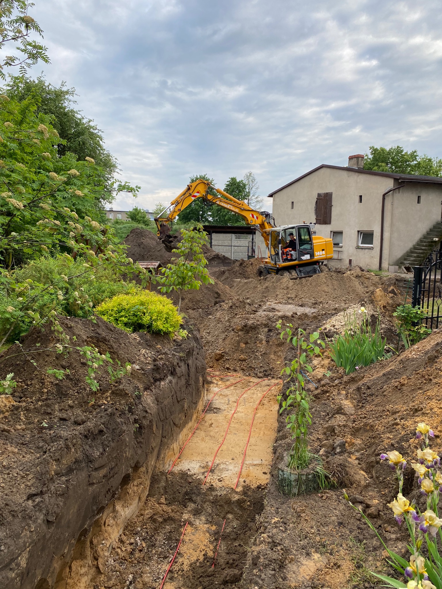 Żółta koparka Liebherr 1404 wykonuje wykop pod instalacje w ogrodzie, widoczne czerwone rury w wykopie oraz operator w kabinie, w tle dom z zamkniętymi okiennicami.