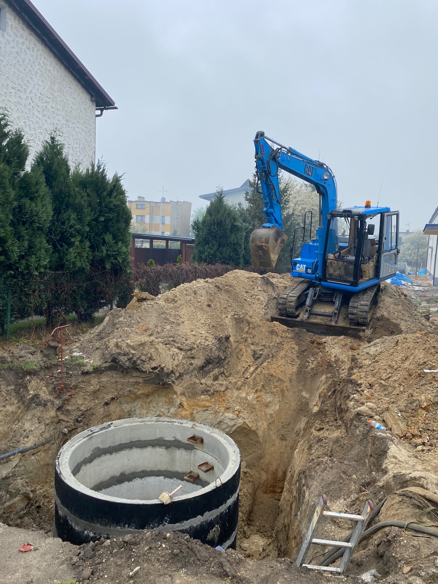Niebieska koparka CAT obok betonowego kręgu w wykopie, drabina oparta o ścianę wykopu, w tle zabudowa mieszkalna w pochmurny dzień.