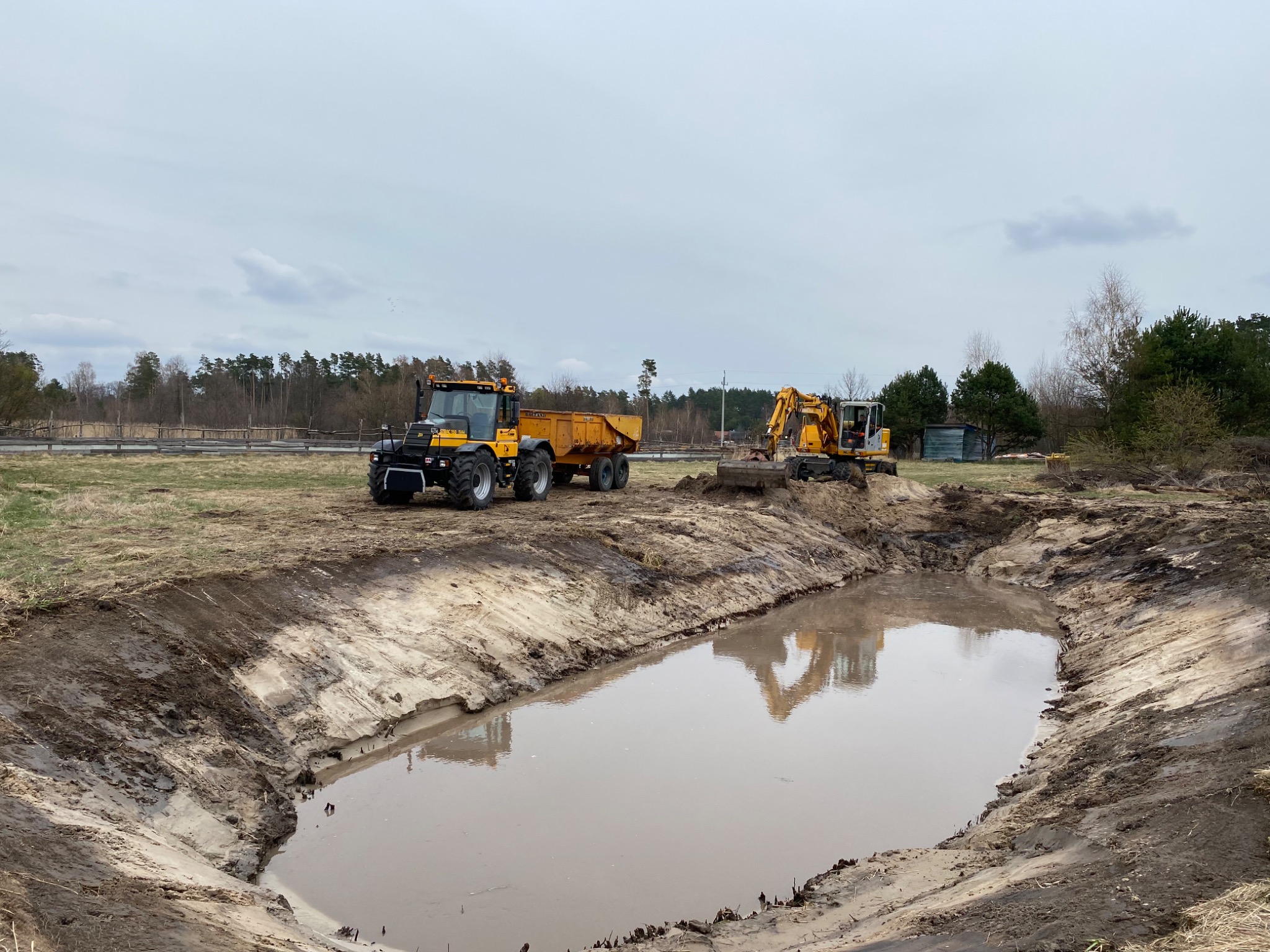 Żółty traktor z przyczepą i koparka pracują przy wykopanym stawie wypełnionym wodą, widoczne ślady prac ziemnych i las w tle.