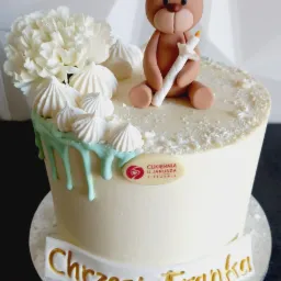 Tort na Chrzest z dekoracją z białych kwiatów, bezików i figurką misia trzymającego świecę, z napisem 'Chrzest Franka' w złotym kolorze.