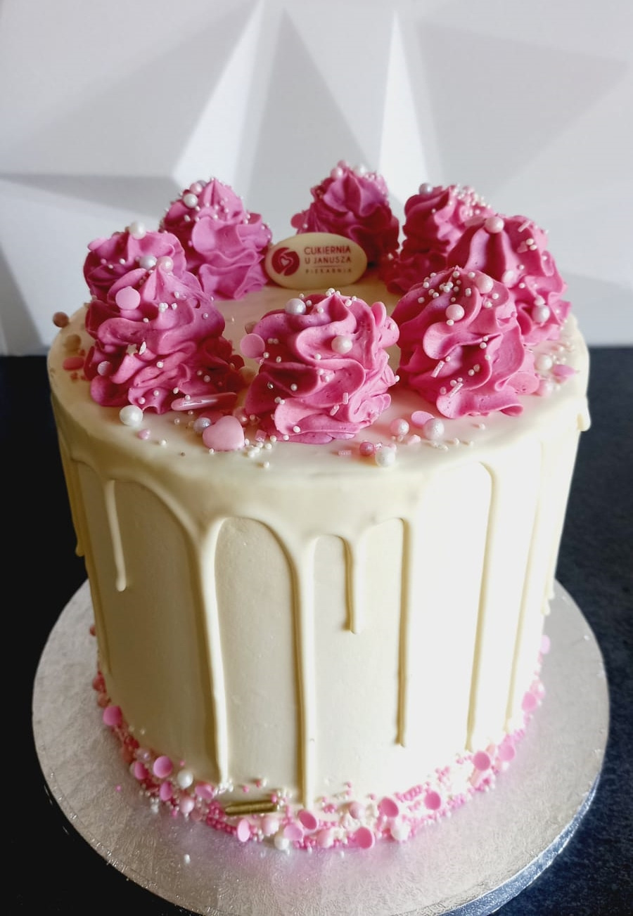 Tort z białym lukrem, ozdobiony różowymi rozetkami z kremu, posypany białymi i różowymi kuleczkami, z logo cukierni na górze.