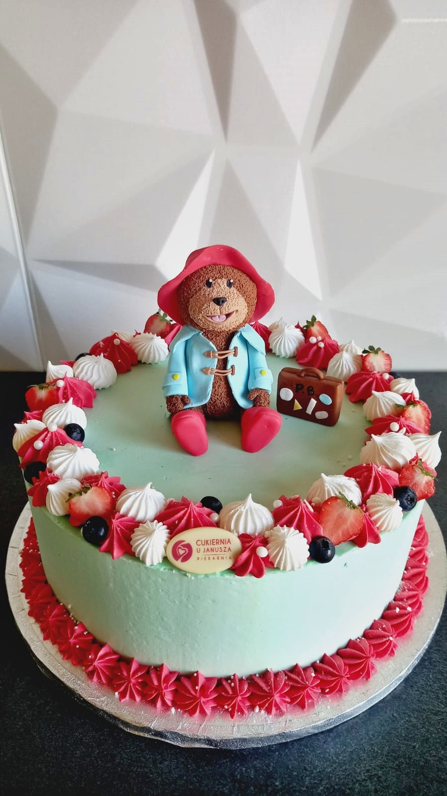 Tort z jasną pistacjową polewą, udekorowany figurką misia Paddingtona z walizką, bezikami, truskawkami i borówkami, z dekoracyjnym rantem z różowej kremówki.
