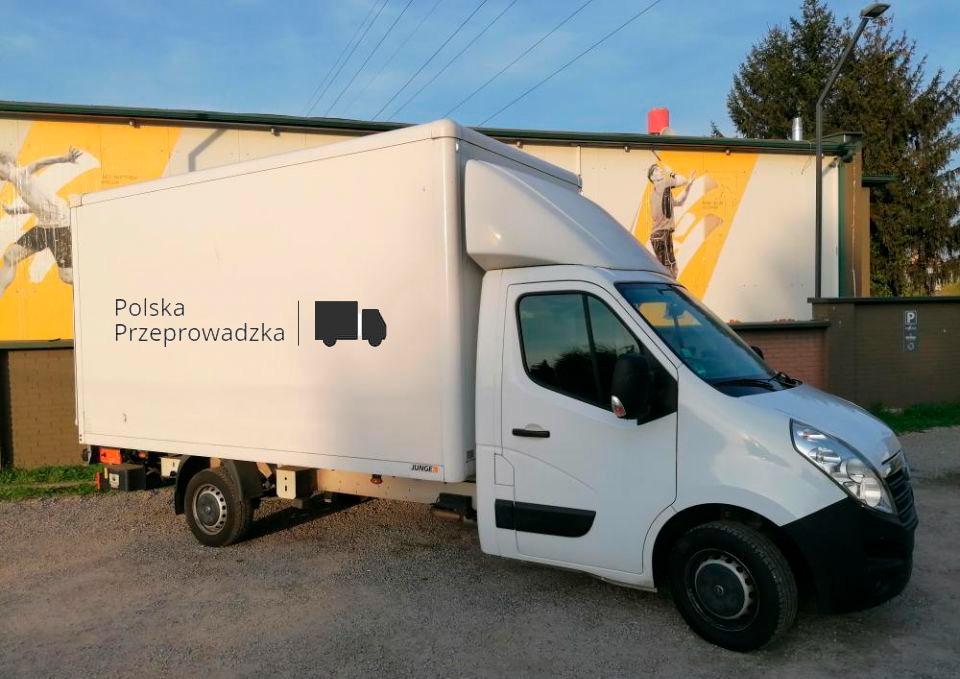 Biały samochód dostawczy z nadrukiem 'Polska Przeprowadzka' i ikoną ciężarówki na boku, zaparkowany przed budynkiem z muralem przedstawiającym osobę malującą.