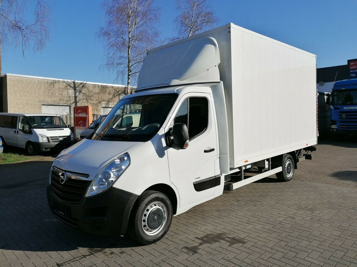 Biały samochód dostawczy Opel z zabudową kontenerową na tle budynku i innych pojazdów, widoczny z przodu pod kątem, w słoneczny dzień.