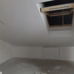 Poddasze po remoncie: świeżo pomalowane ściany na biało, otwarte okno dachowe z drewnianą ramą, widoczny fragment drabiny malarskiej z niebieską taśmą zabezpieczającą, surowa betonowa podłoga.