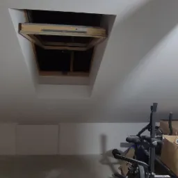 Poddasze po odświeżeniu: białe ściany i sufit, otwarty właz na strych z widoczną konstrukcją drewnianą, widoczne kartony i sprzęt sportowy.