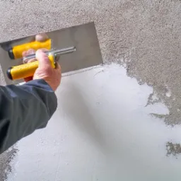 Nakładanie gładzi szpachlowej na ścianę z wyraźną fakturą, widoczna metalowa szpachla z żółtą rączką w dłoni fachowca.