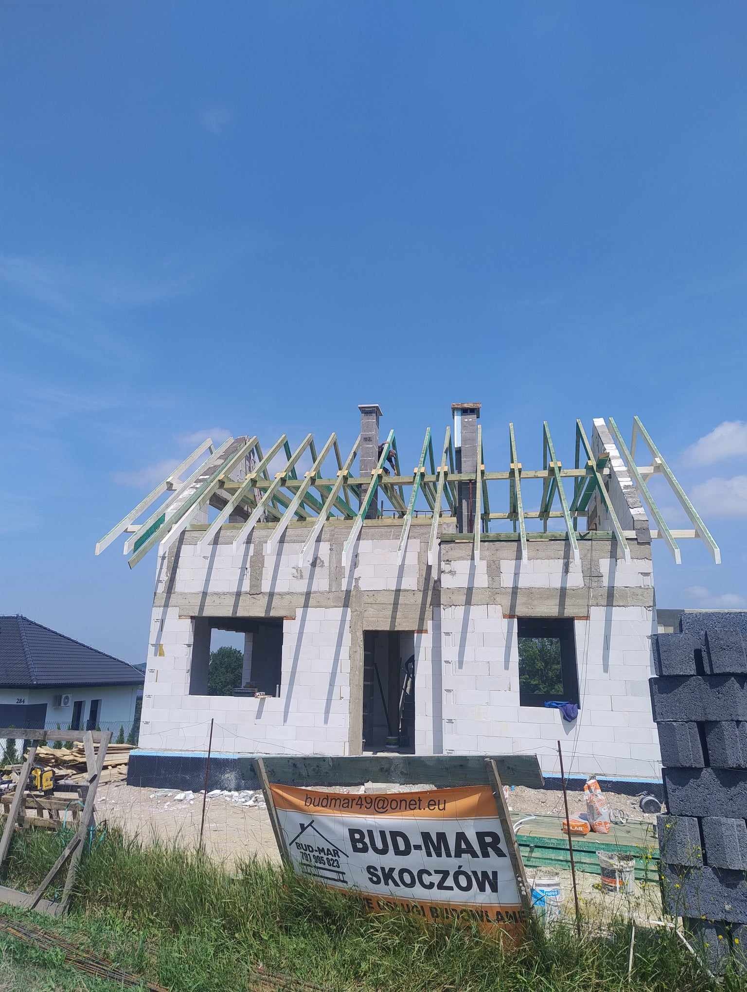 Budowa domu w stanie surowym otwartym: białe ściany z betonu komórkowego, drewniana konstrukcja dachu, widoczny komin i logo firmy budowlanej na ogrodzeniu.