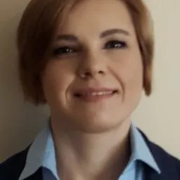 Portret kobiety w niebieskiej koszuli i granatowej marynarce, z krótkimi brązowymi włosami i delikatnym uśmiechem, na neutralnym tle.