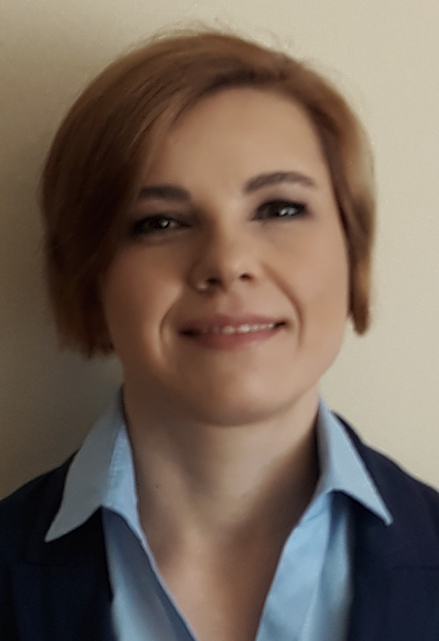 Portret kobiety w niebieskiej koszuli i granatowej marynarce, z krótkimi brązowymi włosami i delikatnym uśmiechem, na neutralnym tle.