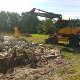 BARMEK - Żółta koparka Volvo EW160 z młotem hydraulicznym kruszy beton, tworząc dużą hałdę pokruszonego materiału i ziemi. W tle drzewa i fragment budynku.