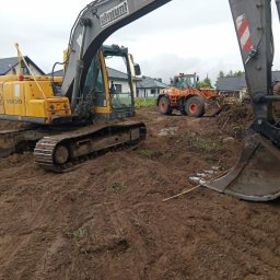 BARMEK - Żółta koparka gąsienicowa Volvo z logo ELGRUNT i pomarańczowa ładowarka kołowa na błotnistym placu budowy, w tle domy jednorodzinne.
