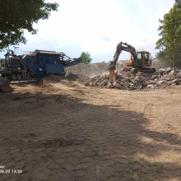 BARMEK - Żółta koparka z młotem hydraulicznym rozbija betonowy gruz, tworząc hałdy materiału do przetworzenia przez niebieską kruszarkę mobilną, widoczną na tle zieleni.