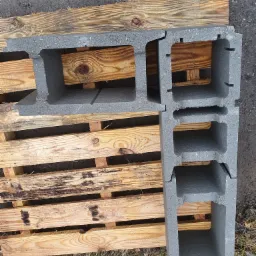 Szare bloczki betonowe ułożone na drewnianej palecie, widoczne pęknięcie na jednym z bloczków, tło stanowi ziemia i trawa.