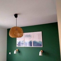 AdRem Jabłoński Adrian - Pomieszczenie z zieloną ścianą, trzema obrazami w białych ramkach, plecioną lampą wiszącą i dwoma kinkietami nad wezgłowiem łóżka.