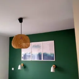 Pomieszczenie z zieloną ścianą, trzema obrazami w białych ramkach, plecioną lampą wiszącą i dwoma kinkietami nad wezgłowiem łóżka.