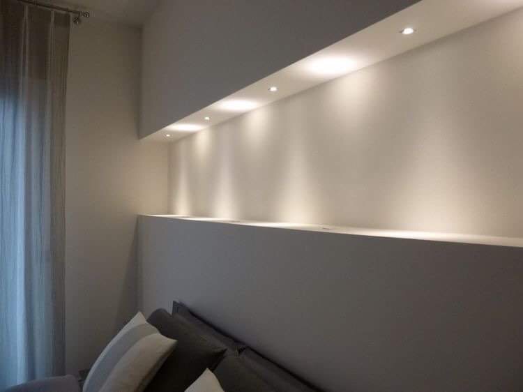 Nowoczesna ściana z wnęką oświetloną punktowymi lampami LED, widok z dołu na poduszki na łóżku, minimalistyczny design wnętrza.