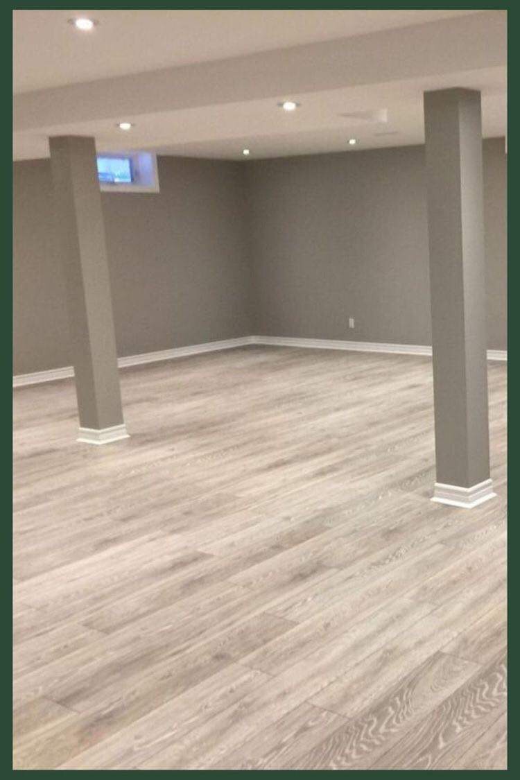 Wykończone wnętrze piwnicy z panelami podłogowymi w odcieniach szarości, pomalowanymi na szaro ścianami, białymi listwami przypodłogowymi i kolumnami nośnymi w kolorze ścian.