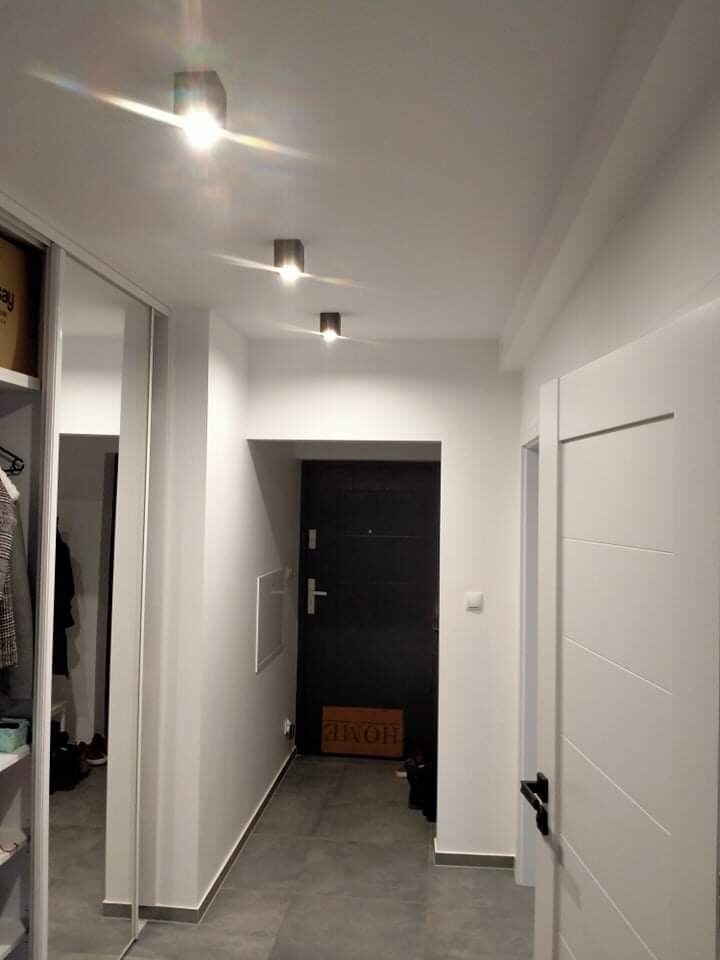 Nowoczesny korytarz z szarą podłogą, białymi ścianami i drzwiami, oświetlony trzema kwadratowymi lampami sufitowymi, z szafą wnękową z lustrzanymi drzwiami po lewej stronie i czarnymi drzwiami...