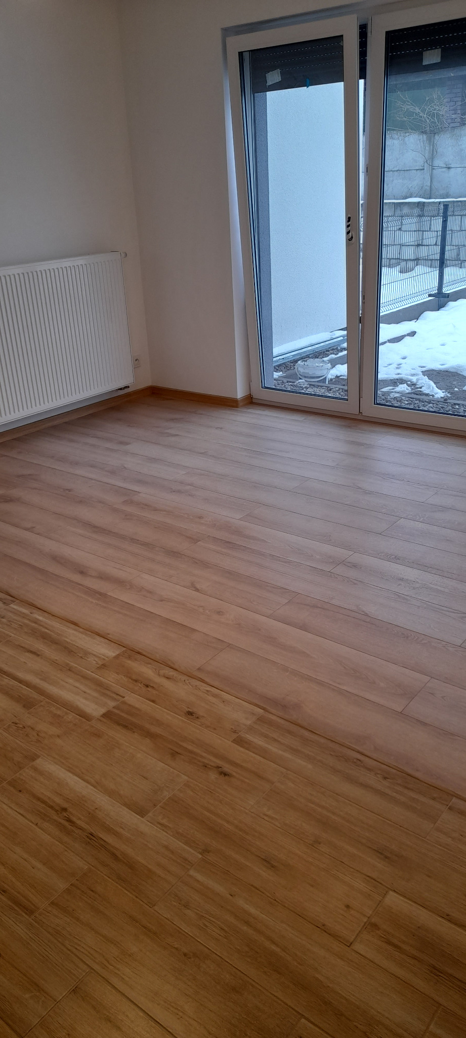 Nowo położona podłoga z paneli w odcieniach brązu, widok na biały grzejnik i przeszklone drzwi balkonowe z widokiem na zaśnieżony teren.