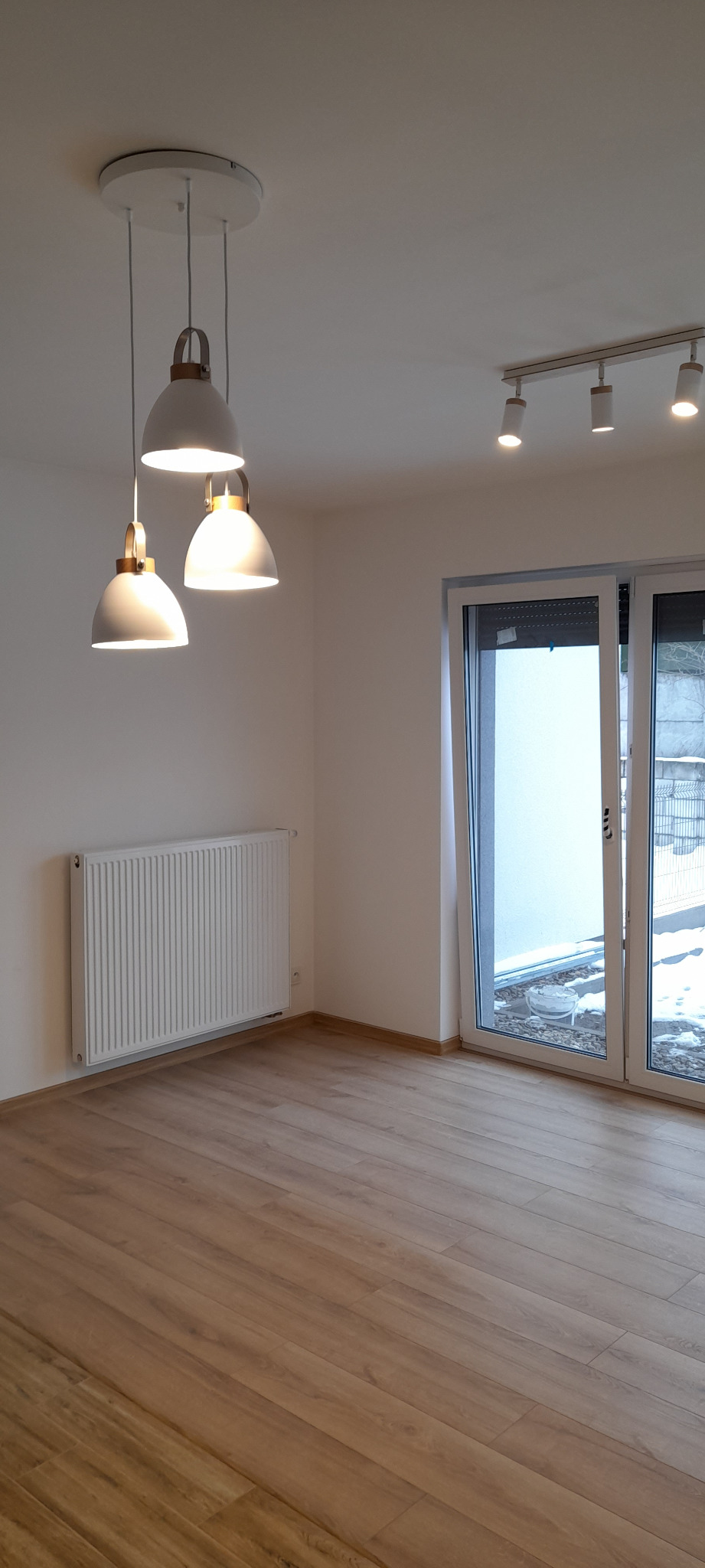 Pusty pokój z parkietem, białymi ścianami, grzejnikiem i trzema wiszącymi lampami z drewnianymi elementami oraz listwą z reflektorami na suficie, z widokiem na zewnątrz przez przeszklone drzwi...
