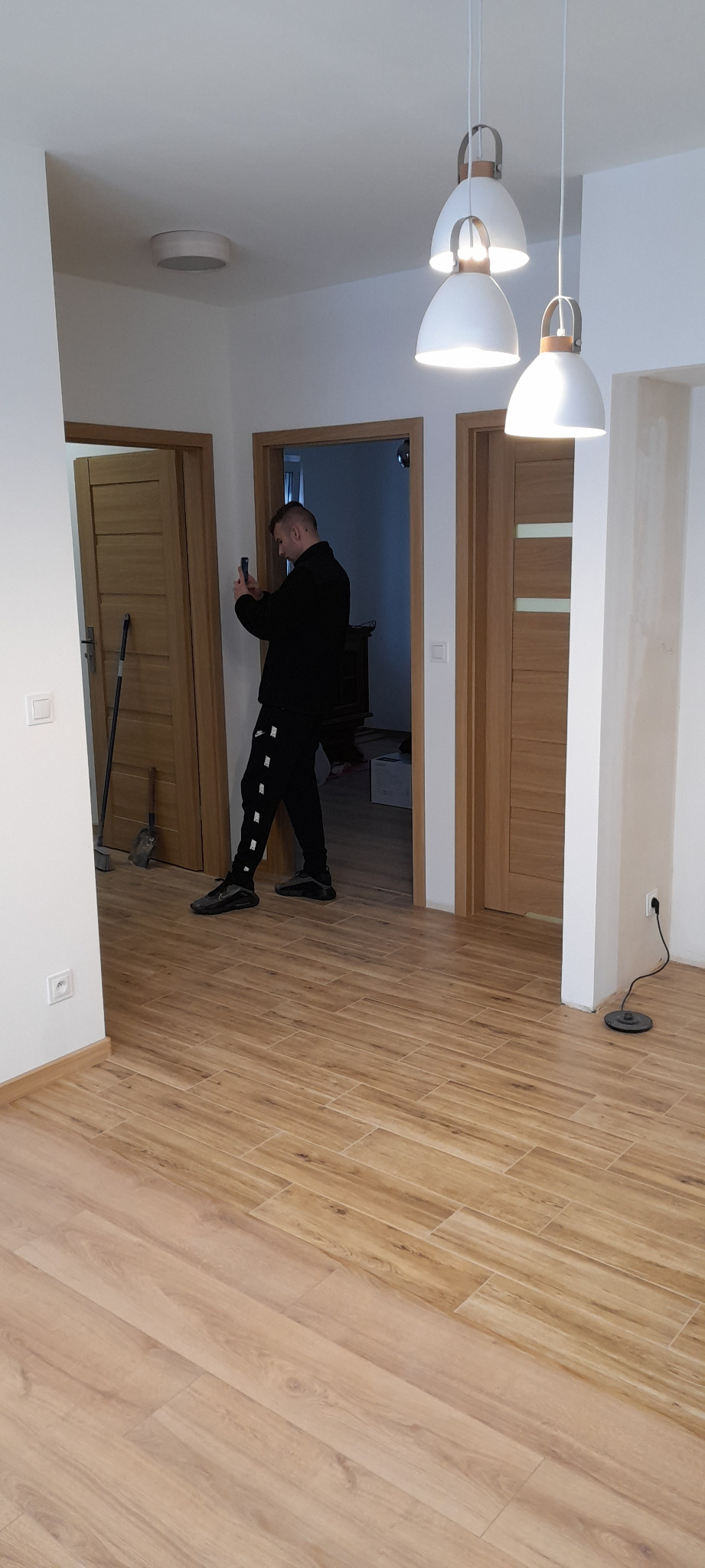 Wnętrze mieszkania po remoncie z drewnopodobną podłogą, trzema wiszącymi lampami i trzema parami drzwi. Mężczyzna w czarnym ubraniu robi zdjęcie telefonem.