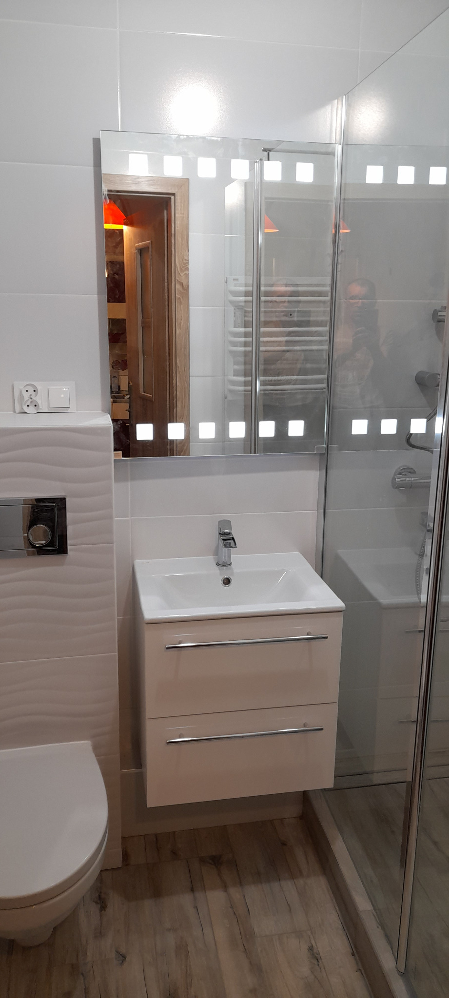 Nowoczesna łazienka z białą umywalką z szafką, podświetlanym lustrem i kabiną prysznicową, widoczne płytki imitujące drewno na podłodze i dekoracyjne płytki na ścianie, odbicie osoby w lustrze.