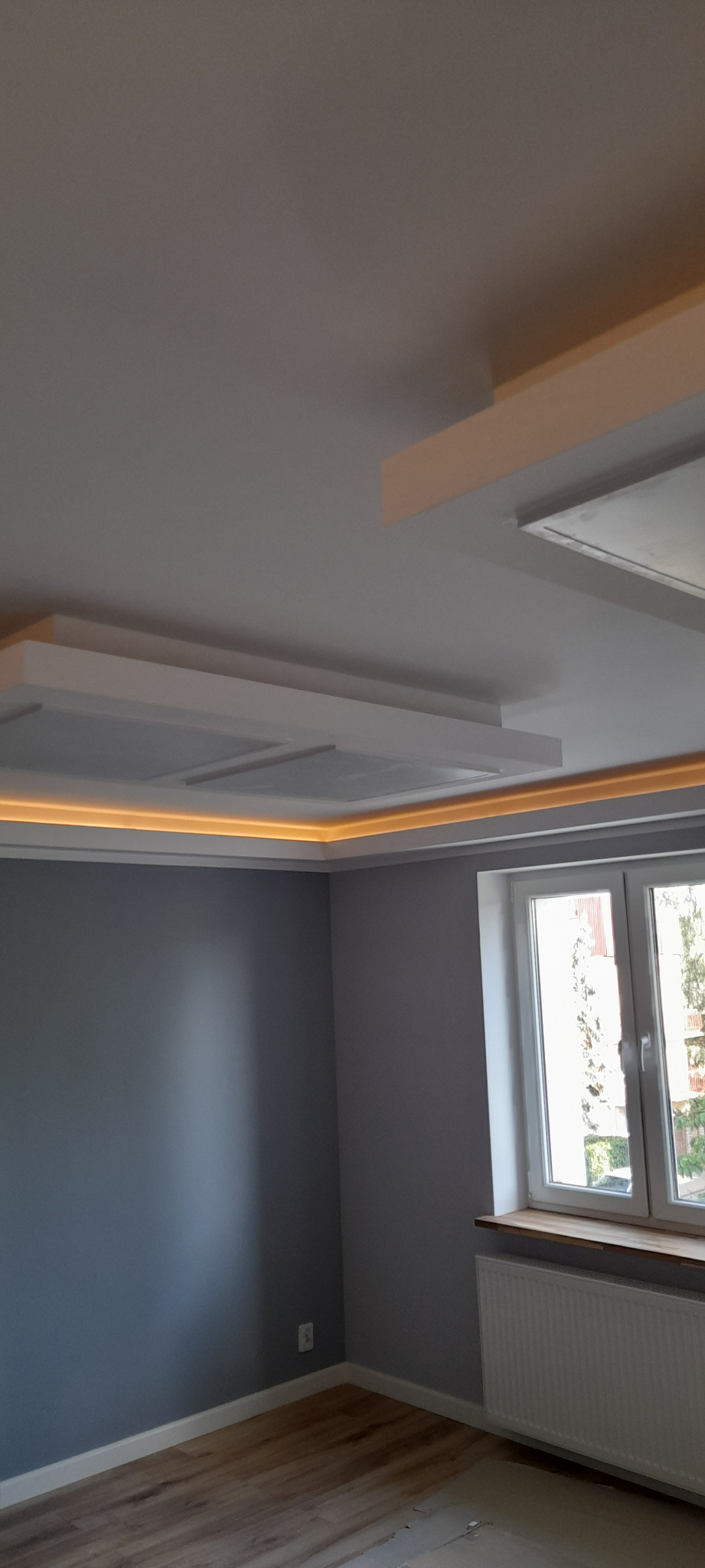 Nowoczesny sufit podwieszany z oświetleniem LED w narożniku szarego pokoju z oknem i grzejnikiem.
