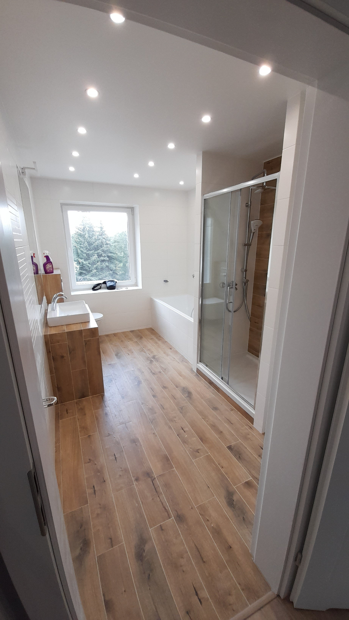 Nowoczesna łazienka po remoncie z drewnopodobną podłogą, kabiną prysznicową z przesuwnymi drzwiami, białą wanną oraz umywalką na szafce z fakturą drewna, oświetlona punktowymi lampami sufitowymi.